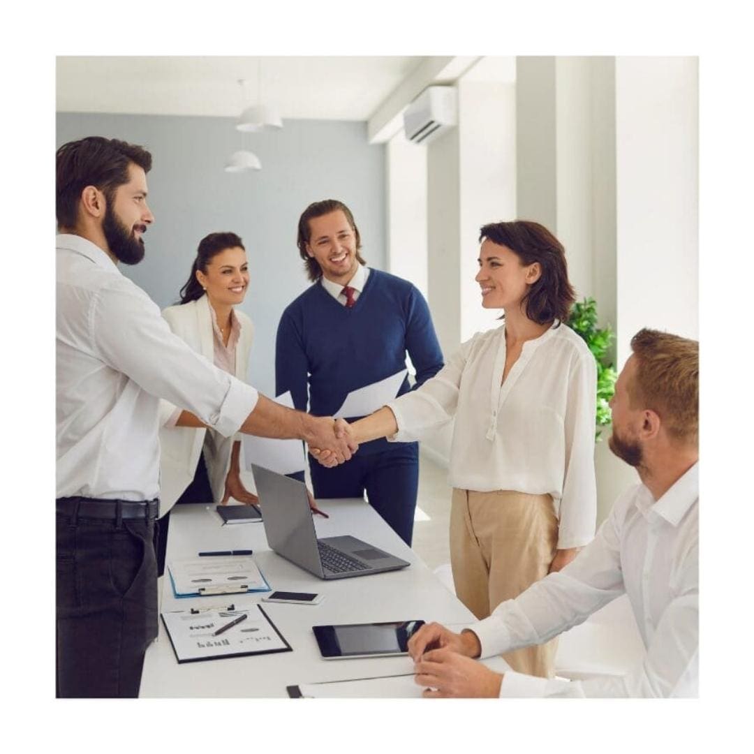 Female professional shakes hands with a male client while the rest of her team stand around the desk.