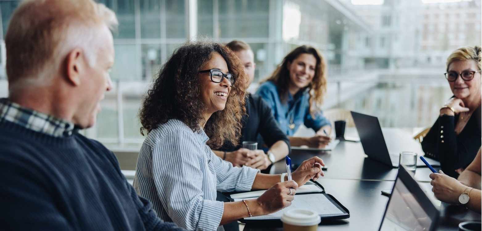 Multiracial adults sit around a conference table with laptops and notepads on hand.